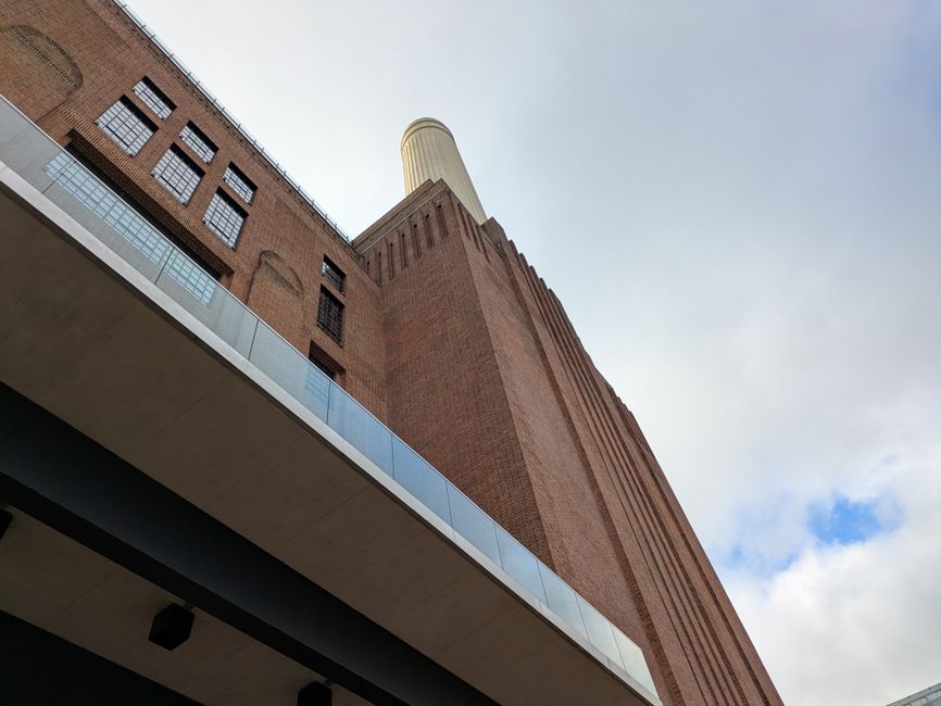 A low-angle view of modern and historic urban architecture in Battersea, showing a curved glass office building on the left with reflective windows, a tall brick chimney in the centre, and a large his